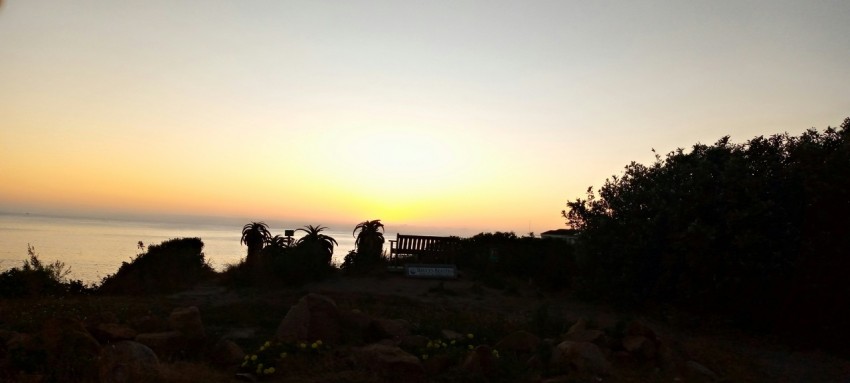 the sun is setting over the ocean with a bench in the foreground