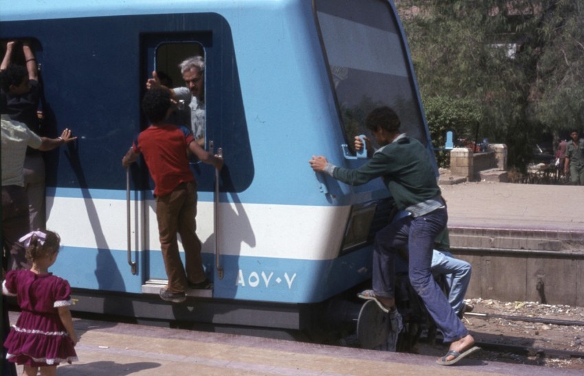 man in green jacket and black pants standing beside blue and white train during daytime