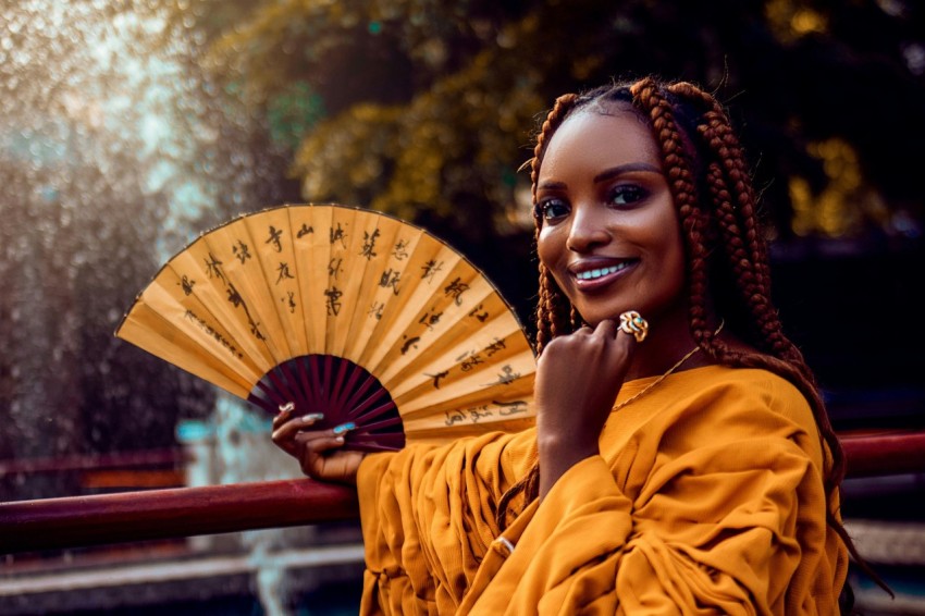 a woman with braids holding a fan in front of a fountain