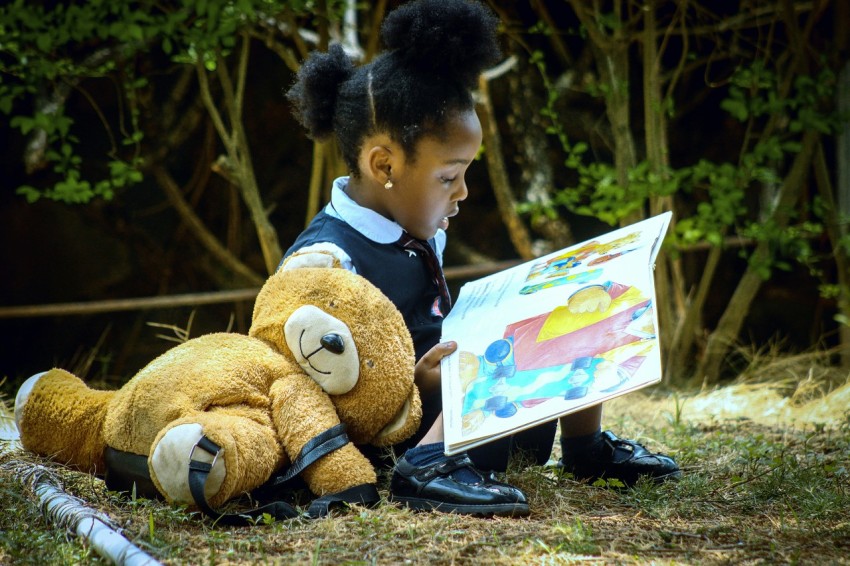 a young girl sitting on the ground reading a book to a teddy bear