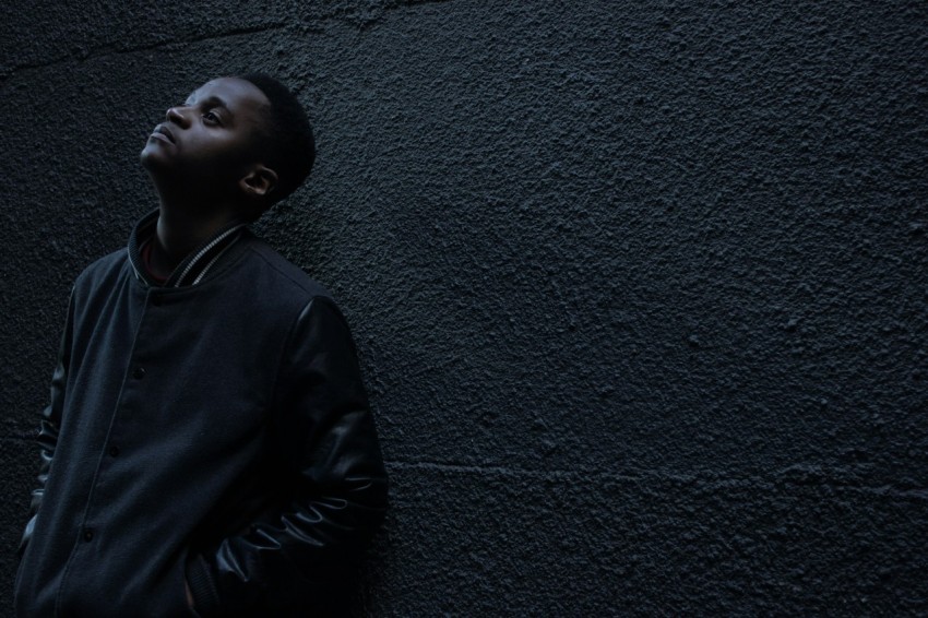 man wearing black button up jacket standing and leaning against the wall while looking up