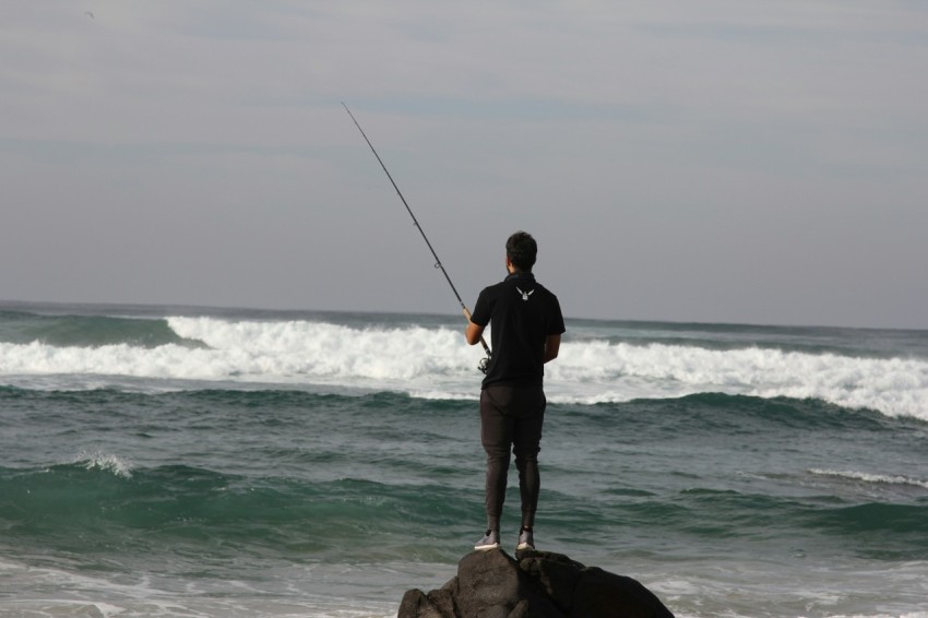 man in black jacket and black shorts holding fishing rod standing on brown rock near sea