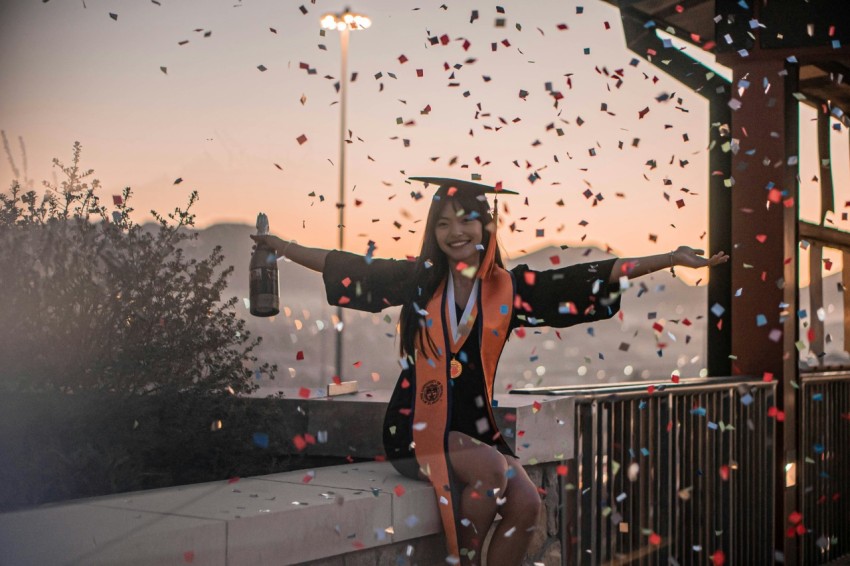 a woman in a graduation cap and gown sitting on a ledge with confetti