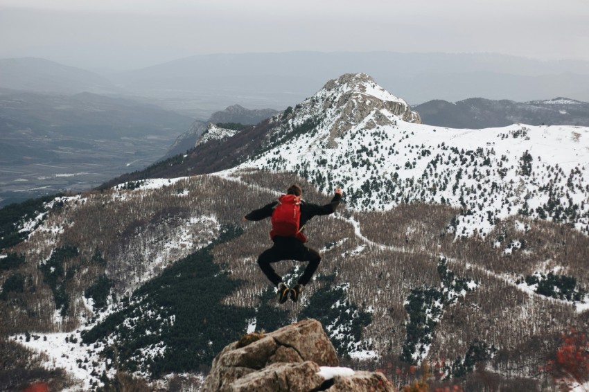 jumping man on rock facing mountains with snow ooS