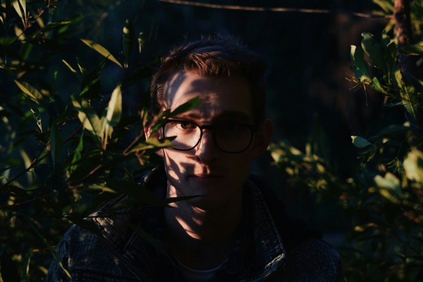 photography of man standing while wearing eyeglasses between near green leaf plants