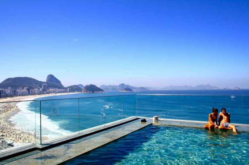 a couple sitting on the edge of a swimming pool overlooking a crowded beach and a deep blue sea