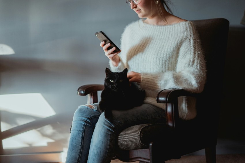 woman in white sweater and blue denim jeans sitting on black leather armchair holding silver iphone p