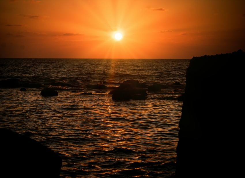 the sun is setting over the ocean with rocks in the foreground