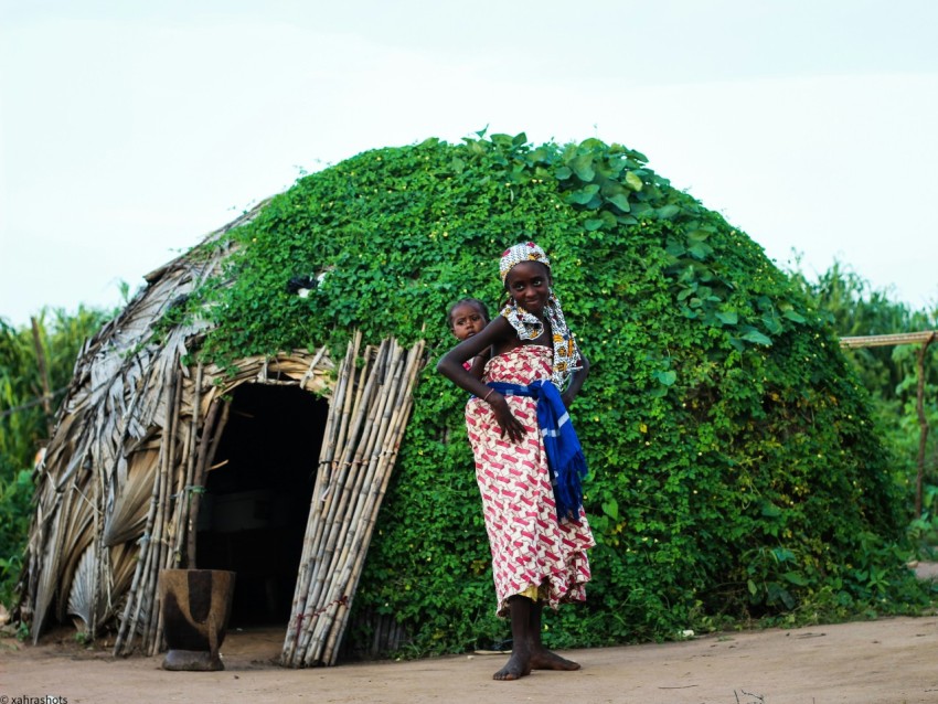 a woman and child standing in front of a hut bx4jZD