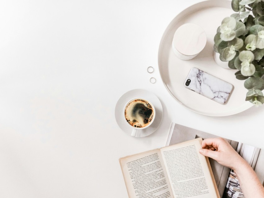 flat lay photography of person holding opened book on tabletop beside cup of black coffee