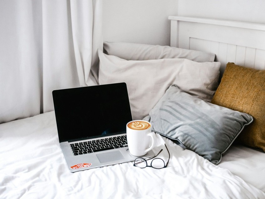 white ceramic mug with coffee on macbook pro near grey pillows on white bed inside bedroom tEQmys5i