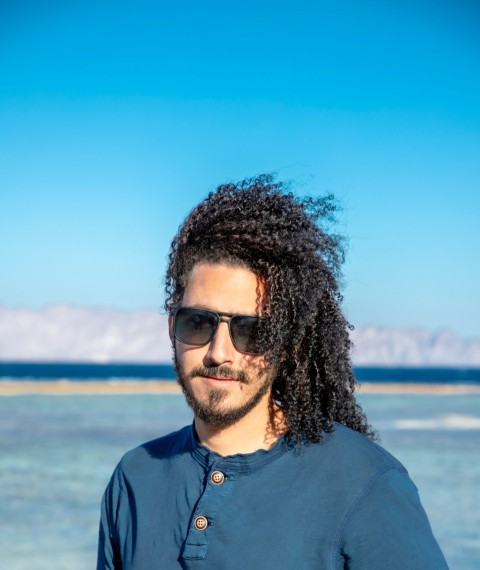 a man with long hair and sunglasses standing in front of a body of water OTn