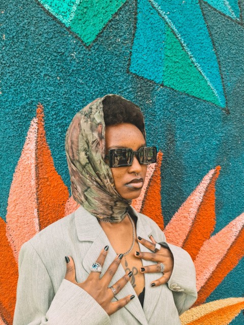 a woman wearing sunglasses and a headscarf stands in front of a colorful wall