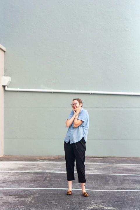 woman in blue dress shirt and black pants standing beside white wall