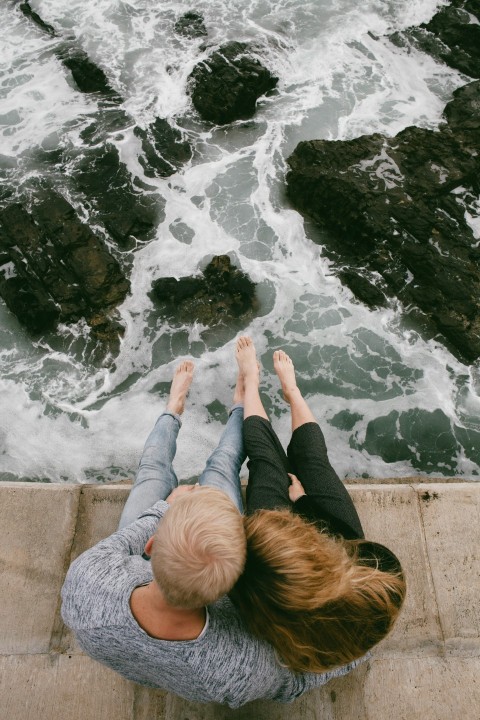 man and woman sitting on cliff near body of water