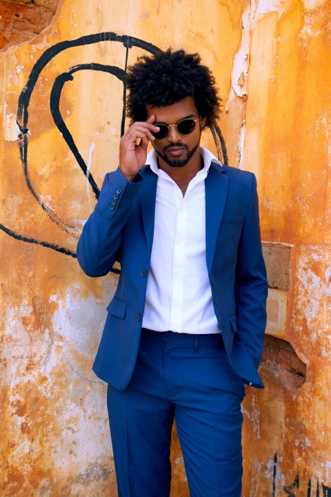 man in blue suit jacket and black sunglasses standing beside brown concrete wall