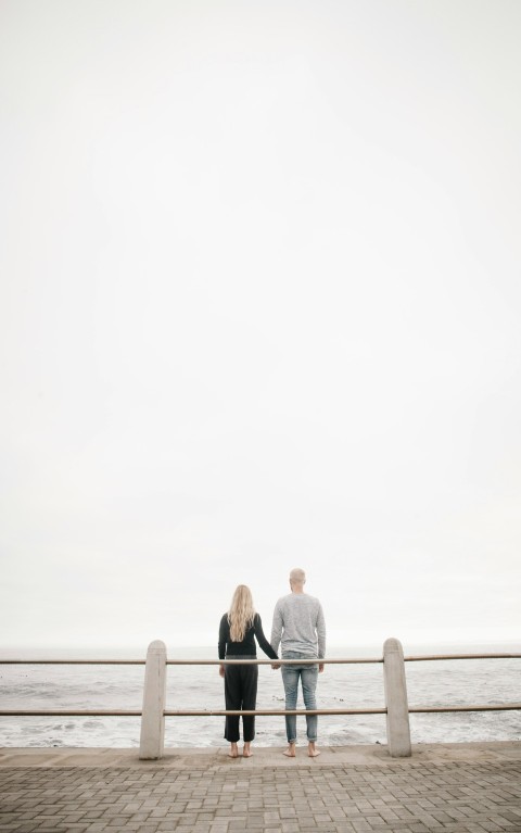 couple leaning on guard rail while holdings hands facing ocean at daytime