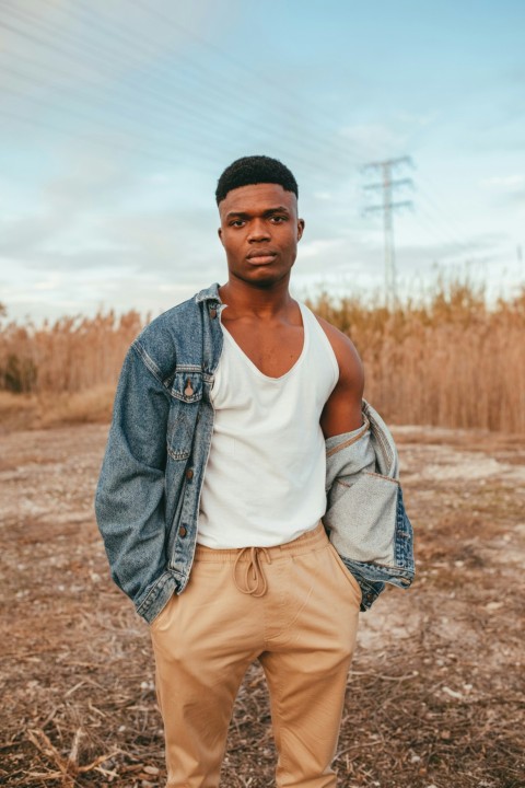 man in white tank top and blue denim jacket standing on brown field during daytime